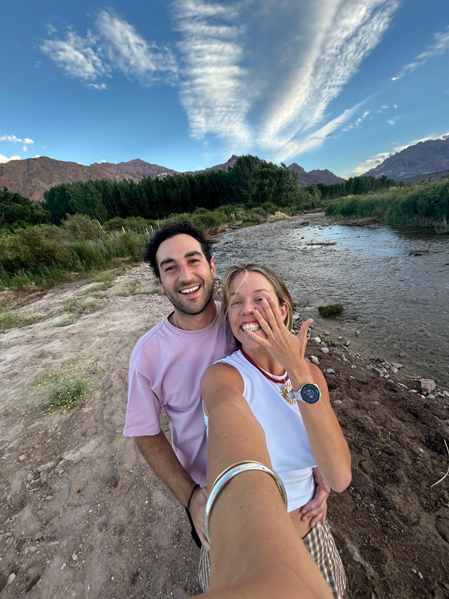 Dante and Cami smiling by a river in the Andean foothills, ring on display